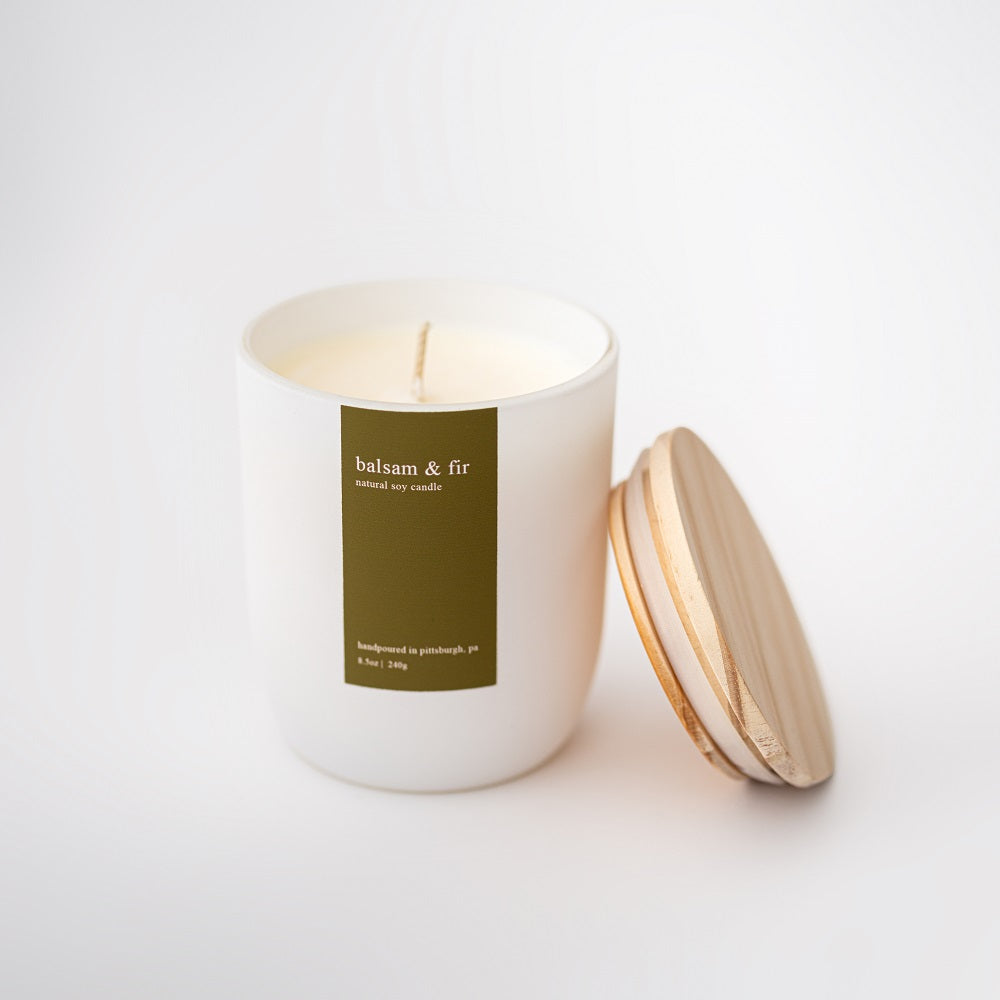 Balsam and fir soy candle featured in its white ceramic jar with wooden lid and green label with white lettering on a white background. 