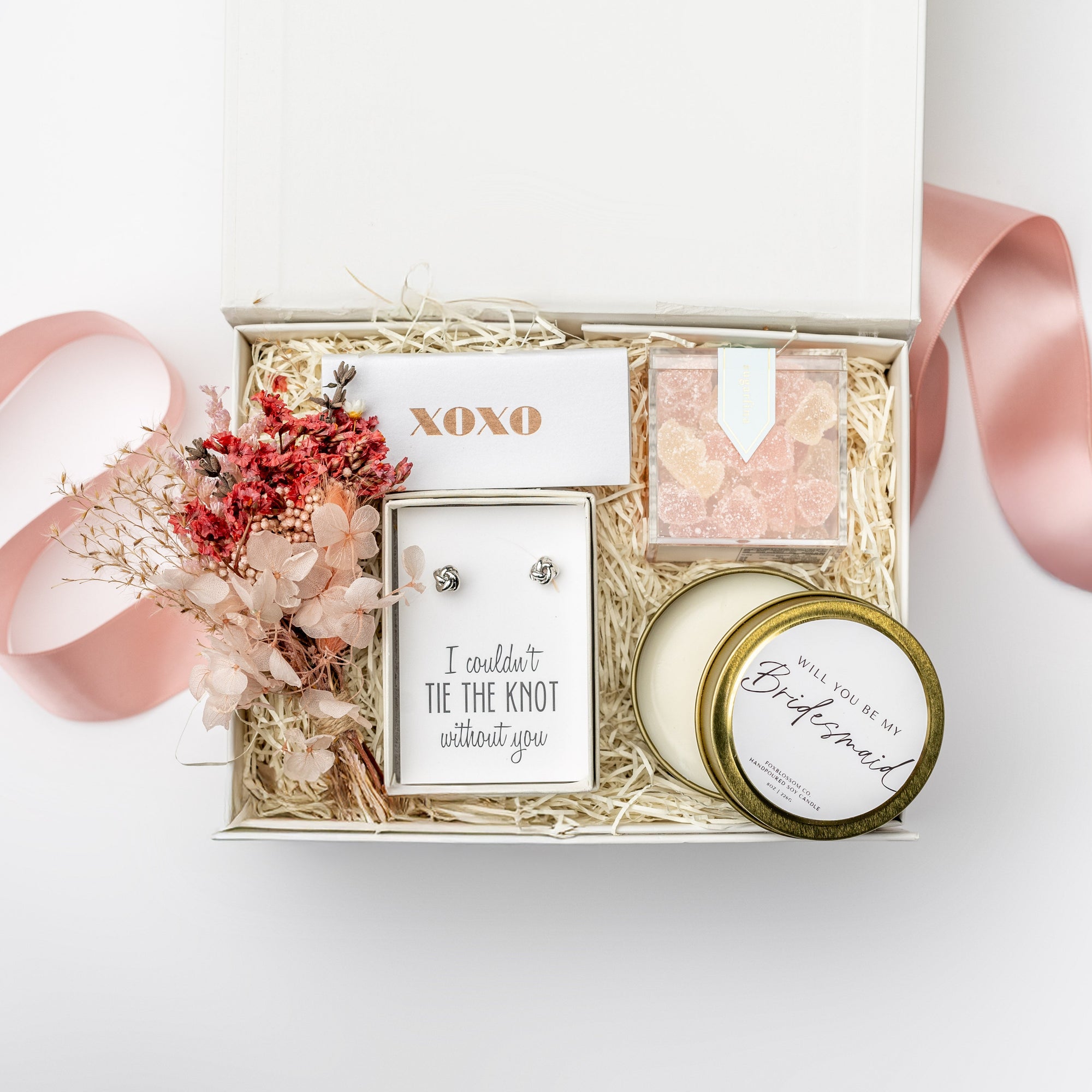 The Be My Bridesmaid Gift Box is featured in our signature white gift box filled with ivory shred. From left to right, it includes a dried floral bundle, a metallic rose gold “XOXO” matchbox in the center, “I Couldn’t Tie the Knot Without You” earrings, champagne bubbly gummy bears, and a “Will You Be My Bridesmaid?” candle in a gold tin. Displayed on a white background with a light pink ribbon surrounding it.