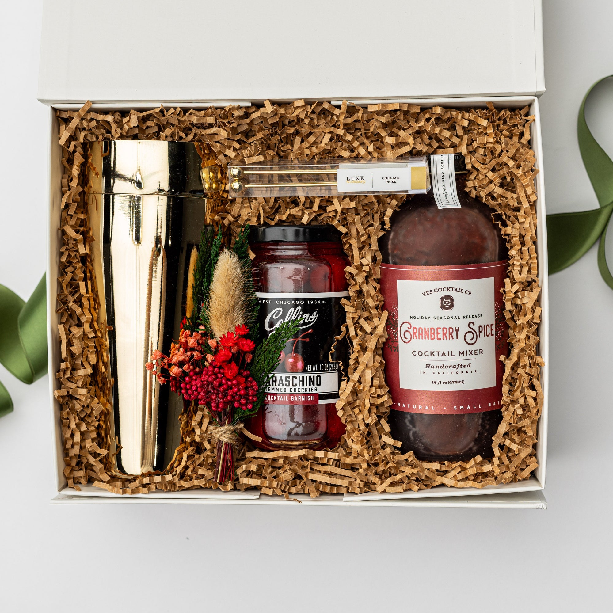 Berry Merry holiday gift box, in a white box with brown shred and a green ribbon underneath it. Inside of the box in the left gold cocktail shaker, in the center set of four gold cocktail picks, stemmed maraschino cherries cocktail garnish , mini floral bouquet, and on the left cranberry spice cocktail mixer red and white label on a white background. 