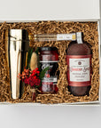 Berry Merry holiday gift box, in a white box with brown shred and a green ribbon underneath it. Inside of the box in the left gold cocktail shaker, in the center set of four gold cocktail picks, stemmed maraschino cherries cocktail garnish , mini floral bouquet, and on the left cranberry spice cocktail mixer red and white label on a white background. 