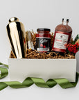 Berry Merry holiday gift box, in a white box with brown shred and a green ribbon around it. Inside of the box in the left gold cocktail shaker, in the center set of four gold cocktail picks, stemmed maraschino cherries cocktail garnish , cranberry spice cocktail mixer red and white label , and on the left mini floral bundle on a white background.