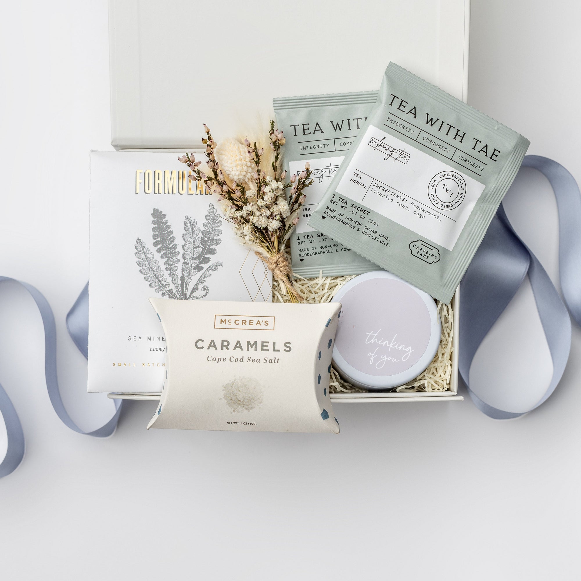 The Better Days Ahead Gift Box is featured in our signature white gift box filled with ivory shred. From left to right, it includes a Formulary Eucalyptus & Peppermint mineral soak, Cape Cod sea salt caramels, a dried floral bundle in the center, and on the right, two calming tea sachets and a “Thinking of You” candle. Displayed on a white background with a light blue ribbon surrounding it.