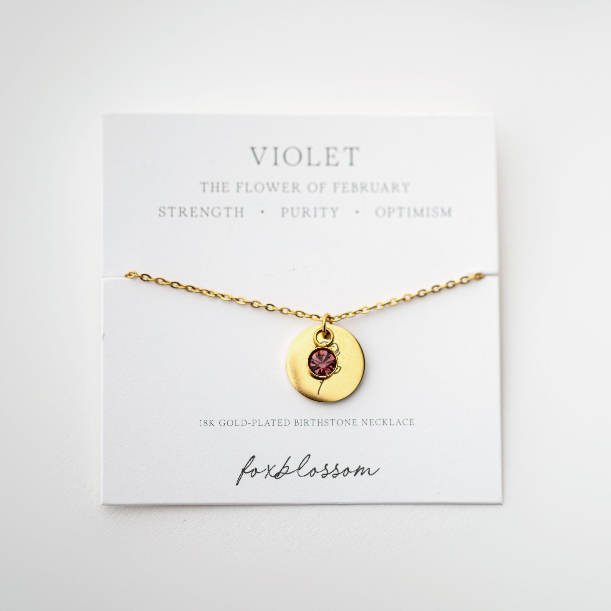 A photograph showcases our delicate gold necklace with a pendant featuring a small violet-colored gemstone and a stylized flower design, presented on our Foxblossom card. 