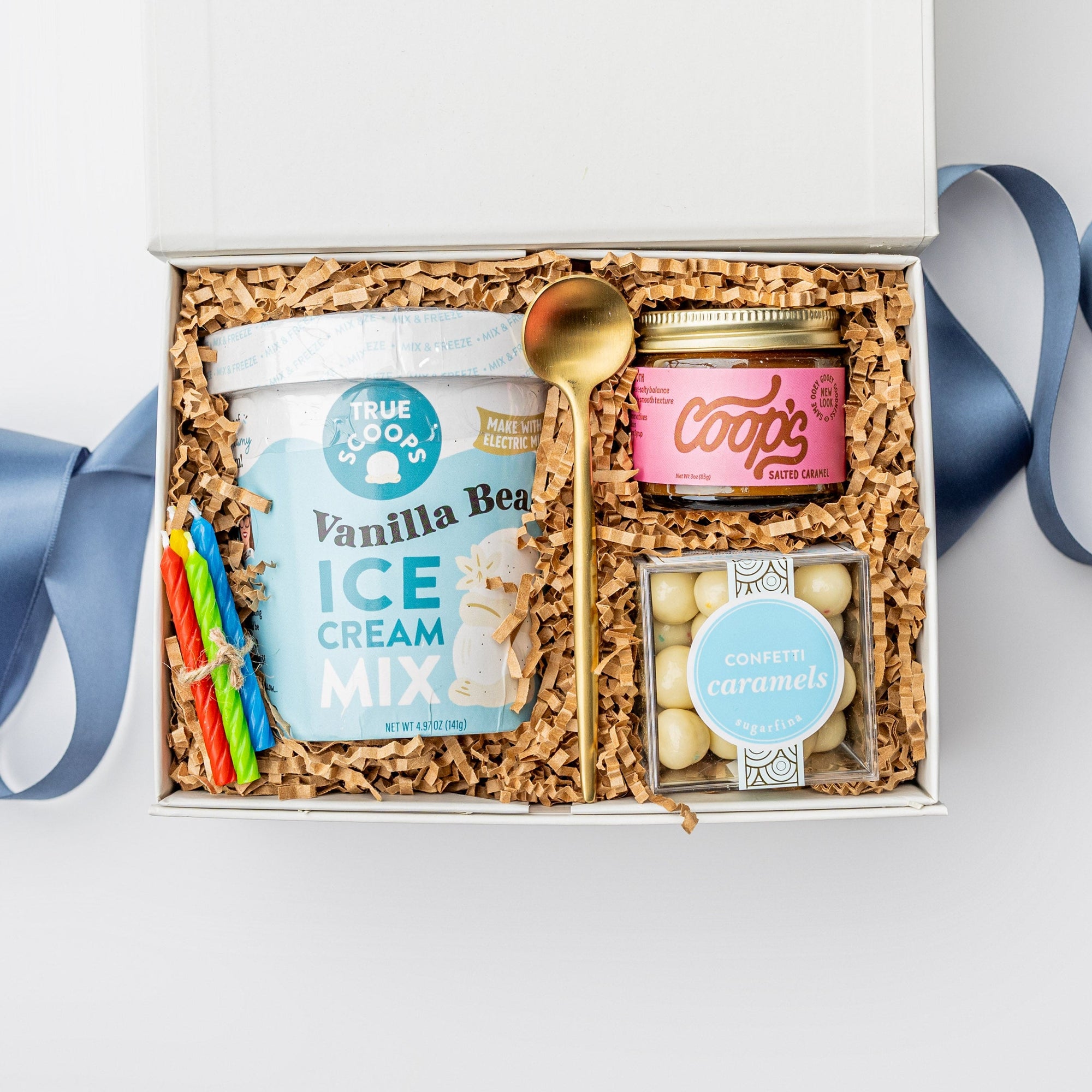 Top view of a white box birthday gift box with brown shreds and a blue ribbon, featuring vanilla ice cream mix, caramel sauce, confetti caramels, a gold spoon, and four mini birthday candles in red, blue, green, and yellow.
