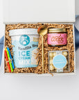 Top view of a white box birthday gift box with brown shreds and a blue ribbon, featuring vanilla ice cream mix, caramel sauce, confetti caramels, a gold spoon, and four mini birthday candles in red, blue, green, and yellow.