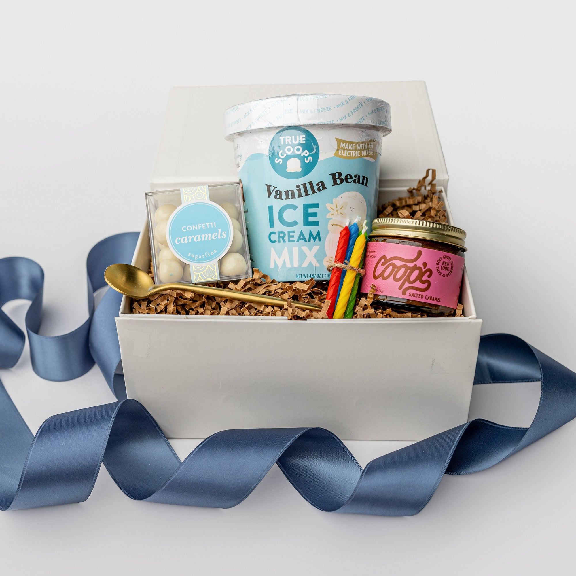 Front view of a white box birthday gift box with brown shreds and a blue ribbon, featuring vanilla ice cream mix, caramel sauce, confetti caramels, a gold spoon, and four mini birthday candles in red, blue, green, and yellow.
