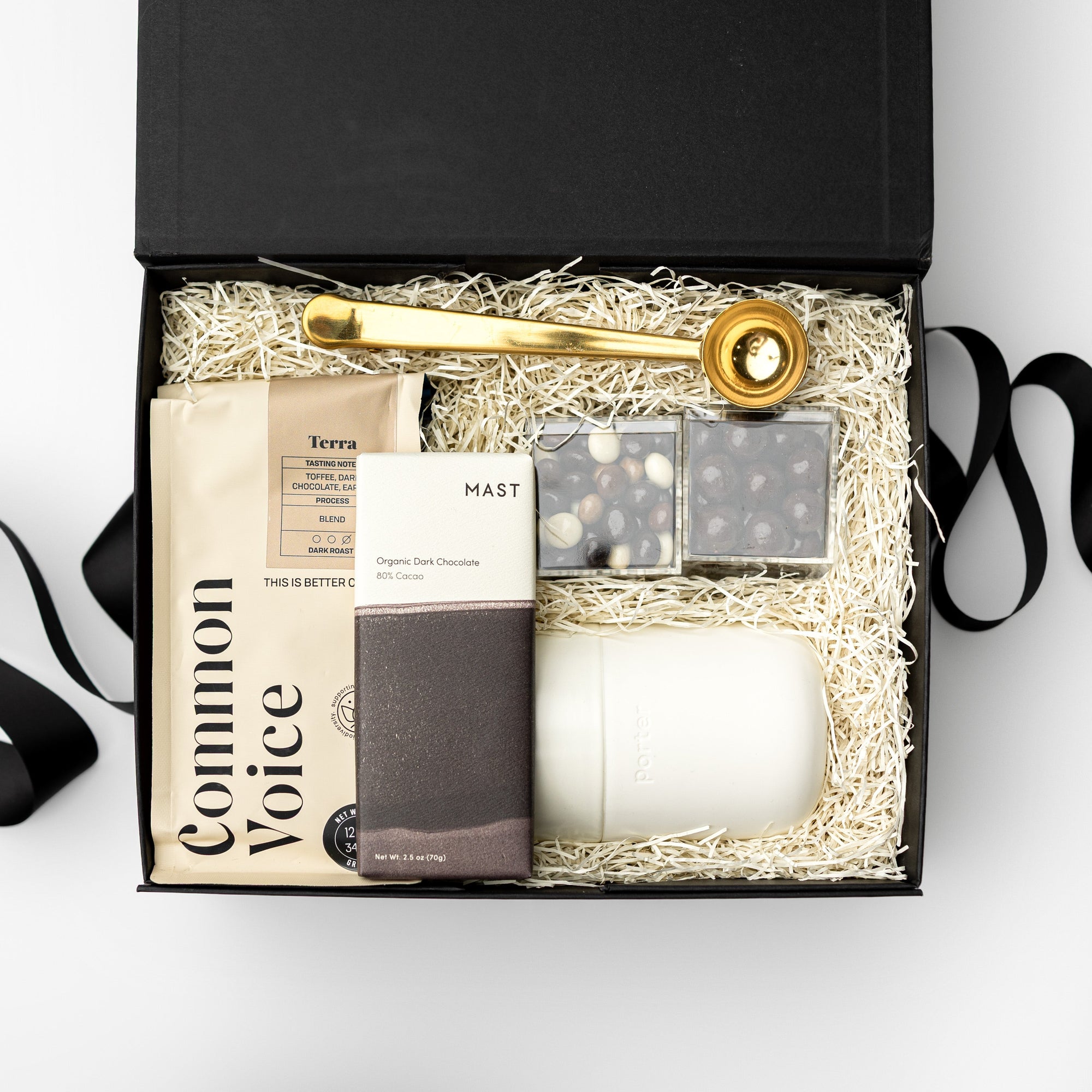 Black gift box with a bag of coffee, dark chocolate bar, two espresso caramels cubes, gold coffee scoop and travel mug, white background