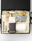 Black gift box with a bag of coffee, dark chocolate bar, two espresso caramels cubes, gold coffee scoop and travel mug, white background