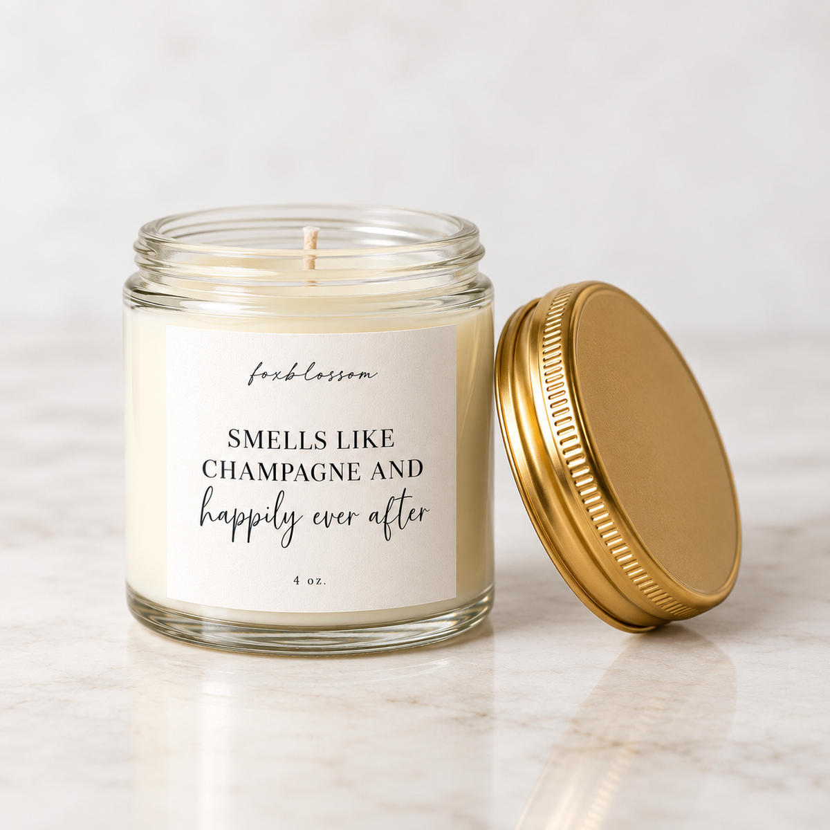 A 4 oz glass jar candle with cream-colored wax and a white label reading 'fkblossom SMELLS LIKE CHAMPAGNE AND happily ever after' in black text. The gold metal lid is placed beside the open jar on a light marble surface.