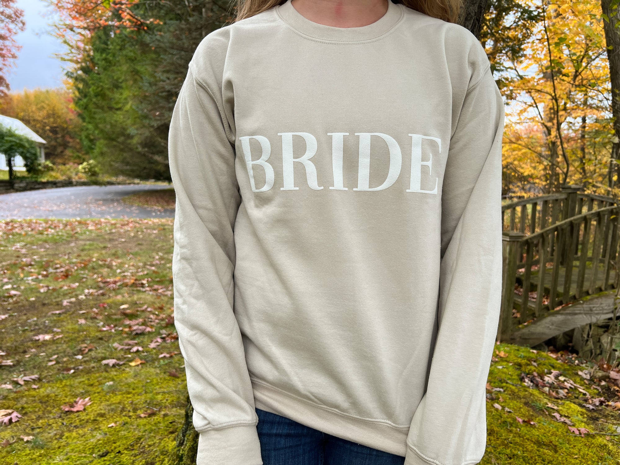 Nude crewneck sweatshirt with large white-embroidered “BRIDE” lettering across the chest worn by a young woman with long hair on a nature.