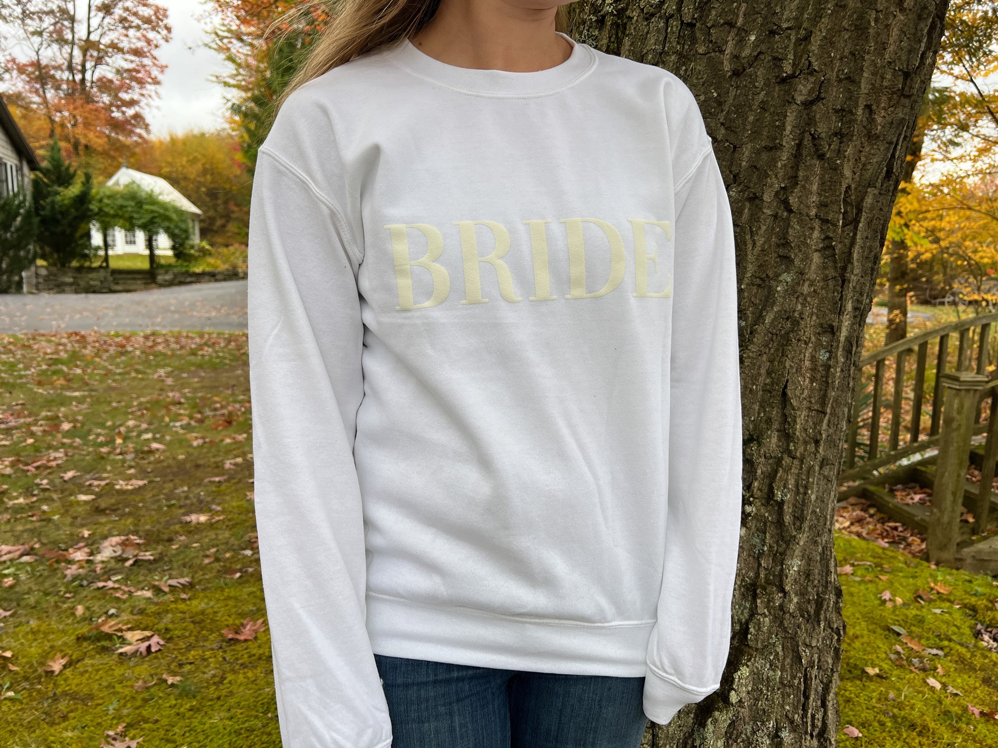 White crewneck sweatshirt with large white-embroidered “BRIDE” lettering across the chest worn by a young woman with long hair on a nature.
