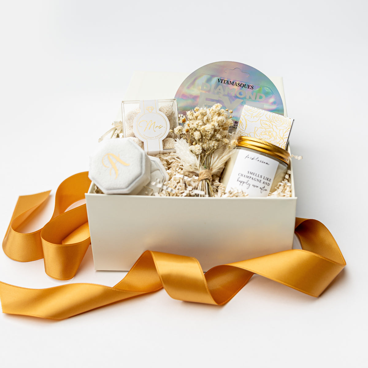 A curated gift box set on a white background, featuring a white box filled with dried flowers, a gold candle jar, a white soap or wax melt, a small clear box with a round monogrammed lid, and an iridescent round item. A gold satin ribbon is draped in front of the box.