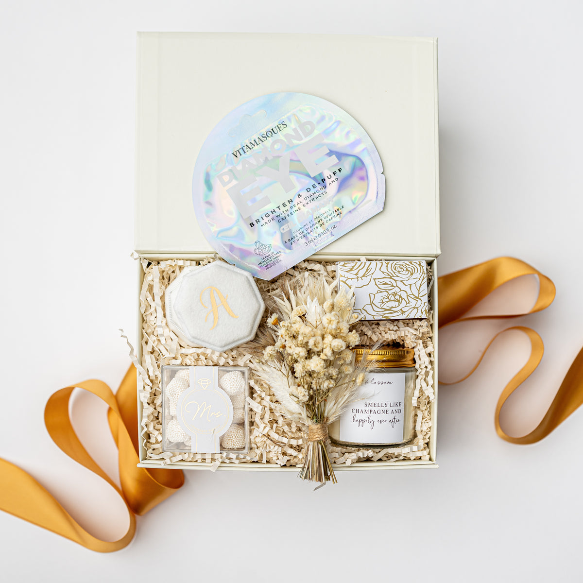 A curated gift box set from Vitamins by Nature on a clean white background. The open cream-colored box is filled with shredded paper and contains a round iridescent sticker, a white wax seal with a gold monogram, a small square soap or wax melt, a dried flower sprig, and a small glass jar candle.