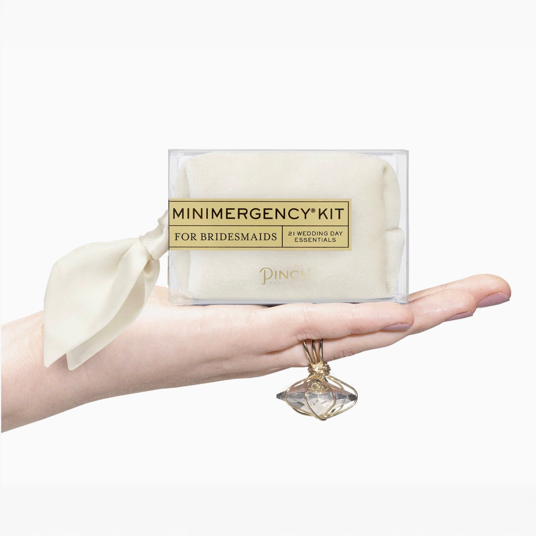 A hand holds a clear plastic package labeled 'MINIMERGENCY KIT FOR BRIDESMAIDS' with a cream ribbon bow. The package contains a folded cream-colored item, and the brand name 'PINCH' is visible on the front.