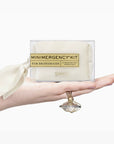 A hand holds a clear plastic package labeled 'MINIMERGENCY KIT FOR BRIDESMAIDS' with a cream ribbon bow. The package contains a folded cream-colored item, and the brand name 'PINCH' is visible on the front.