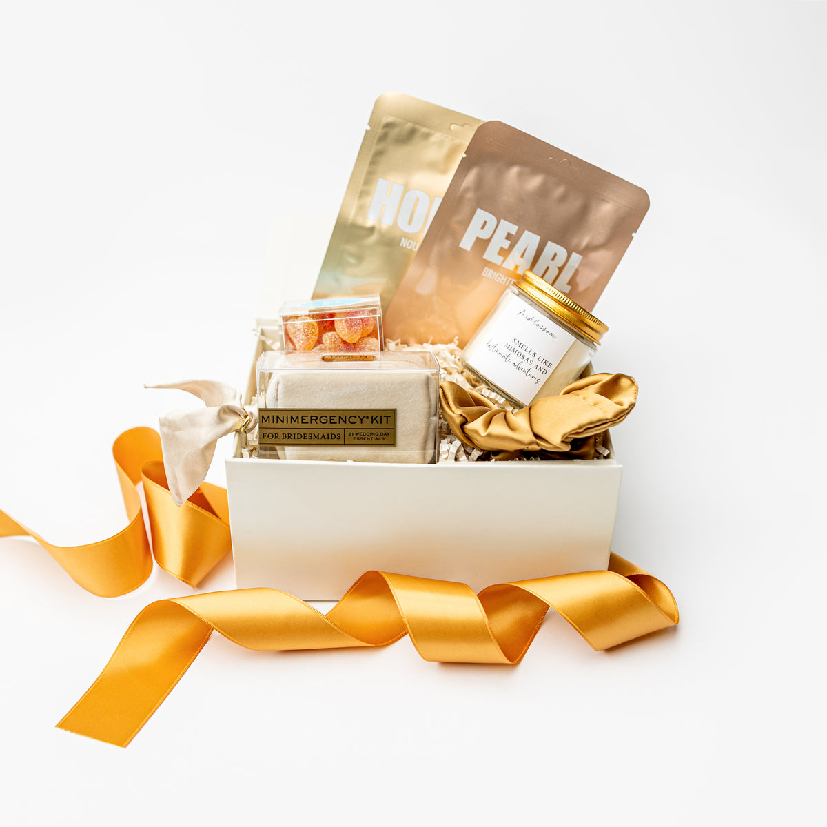 A curated gift box set on a white background, featuring a white box filled with a mini emergency kit, a jar candle, a gold sachet labeled 'HOPE', a brown pouch labeled 'PEARL', and a gold satin ribbon.