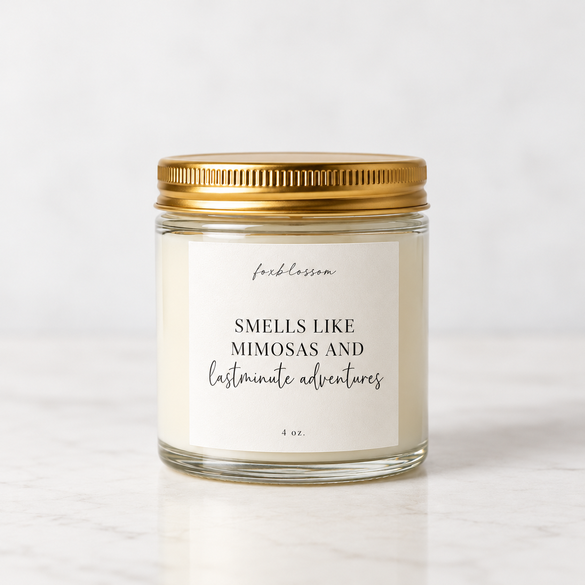 A 4 oz glass jar candle with a gold metal lid and a white label that reads 'feelsblossom' and 'SMELLS LIKE MIMOSAS AND lastminute adventures' on a light marble surface.