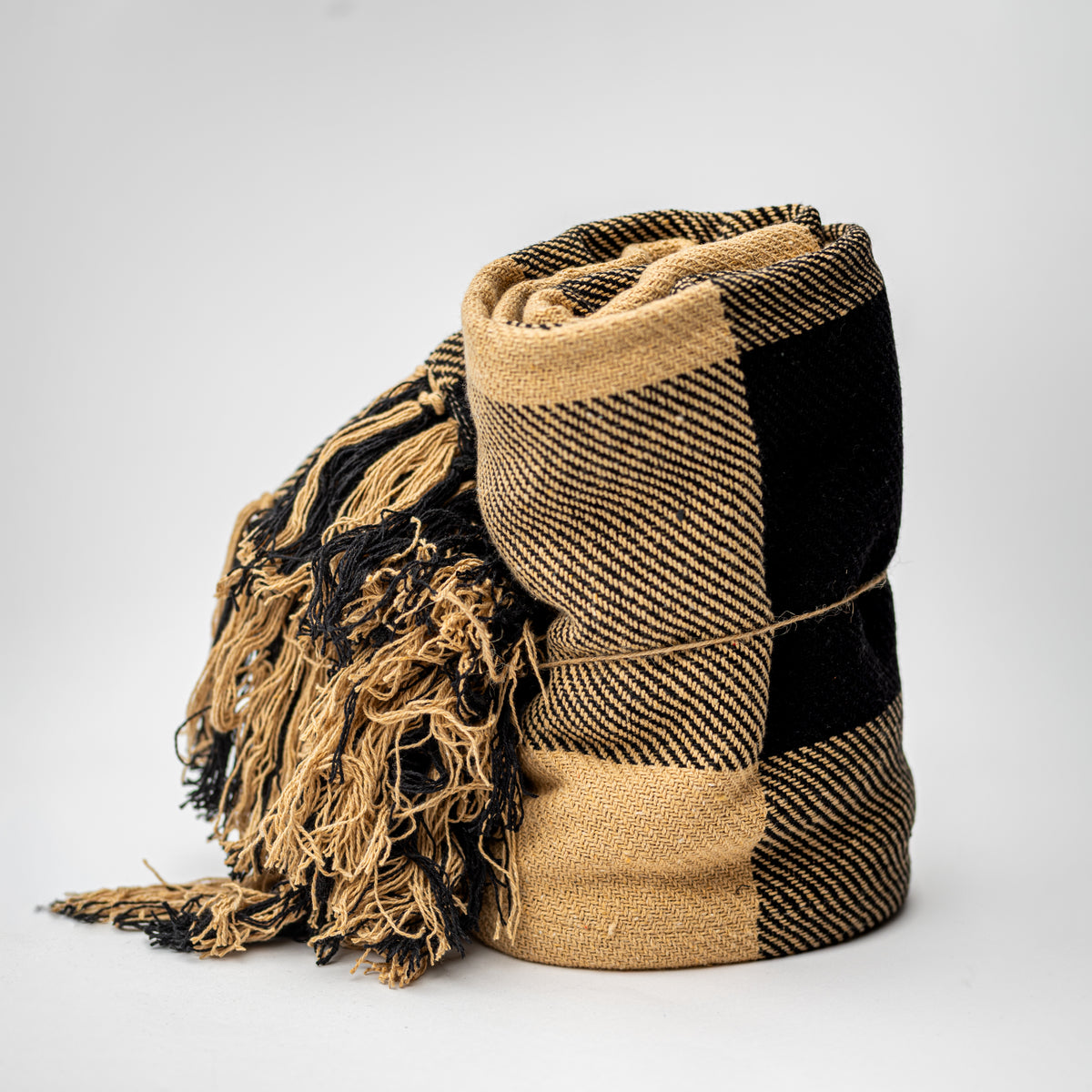 A rolled woven throw blanket with black and tan plaid pattern and fringed tassels, photographed on a light gray studio background.