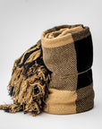 A rolled woven throw blanket with black and tan plaid pattern and fringed tassels, photographed on a light gray studio background.