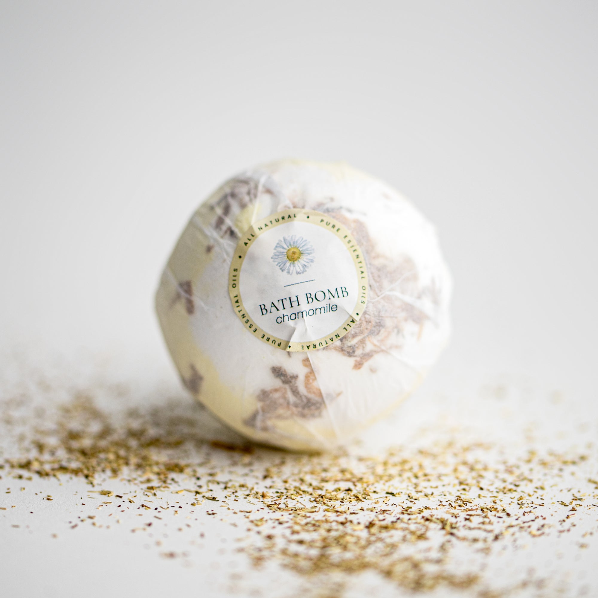 Chamomile bath bomb ,round, in its transparent covering and label that has a chamomile flower and black lettering.  Surrounded by chamomile pieces. 