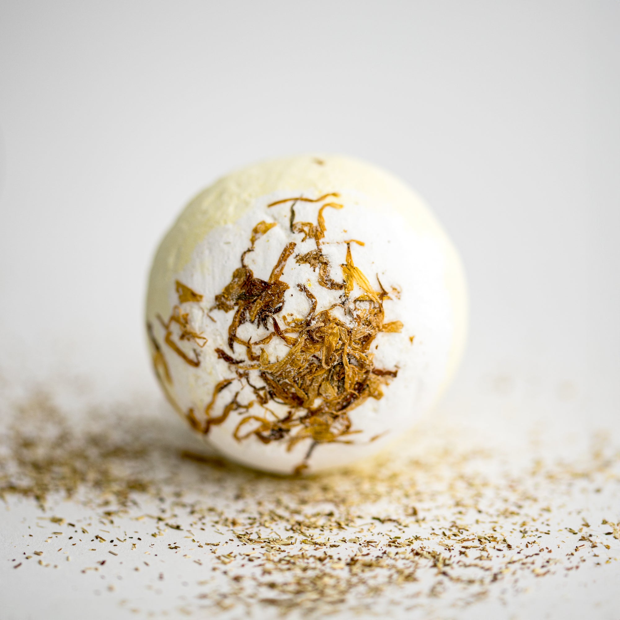 Chamomile bath bomb in its round shape with chamomile pieces around it on a white background. 