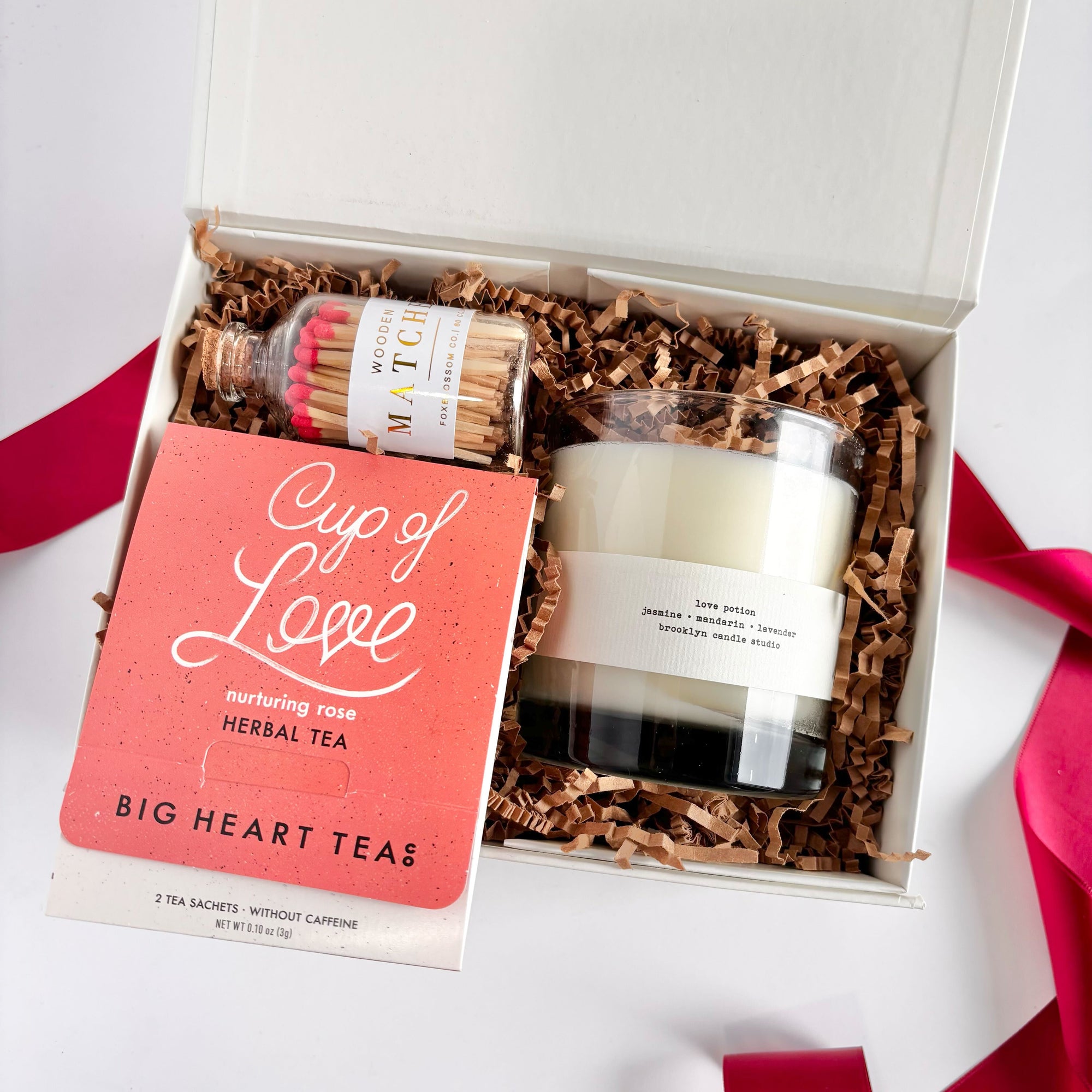 White gift box lined with brown crinkle paper containing a small coral-pink package of Big Heart Tea ‘Cup of Love’ herbal tea sachets, a glass jar of wooden matches with red tips, and the love potion white scented candle in a clear glass holder. 