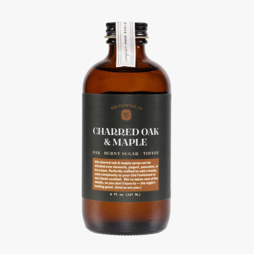Showcasing the Charred Oak & Maple syrup in its dark brown color with a label in dark green almost teal color with white and light brown lettering. Bottle has a black cap sealed with a white sticker to ensure bottle has not been tampered with. 