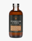 Showcasing the Charred Oak & Maple syrup in its dark brown color with a label in dark green almost teal color with white and light brown lettering. Bottle has a black cap sealed with a white sticker to ensure bottle has not been tampered with. 