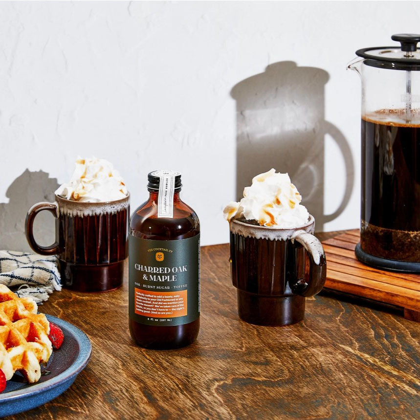 ShowcasIng,  on a dark wooden surface against a plain white wall a french press onto the wall, In the foreground, there are two mugs, each containing a coffee drink topped with whipped cream and drizzled caramel. A bottle of "Charred Oak & Maple" syrup sits between the mugs.  On the left, a plate with waffles and strawberries on a blue plate.  The mugs contain dark brown coffee, topped with  whipped cream and a drizzle of syrup. The syrup bottle is made of dark brown glass with a green and white label.
