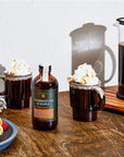 ShowcasIng,  on a dark wooden surface against a plain white wall a french press onto the wall, In the foreground, there are two mugs, each containing a coffee drink topped with whipped cream and drizzled caramel. A bottle of "Charred Oak & Maple" syrup sits between the mugs.  On the left, a plate with waffles and strawberries on a blue plate.  The mugs contain dark brown coffee, topped with  whipped cream and a drizzle of syrup. The syrup bottle is made of dark brown glass with a green and white label.