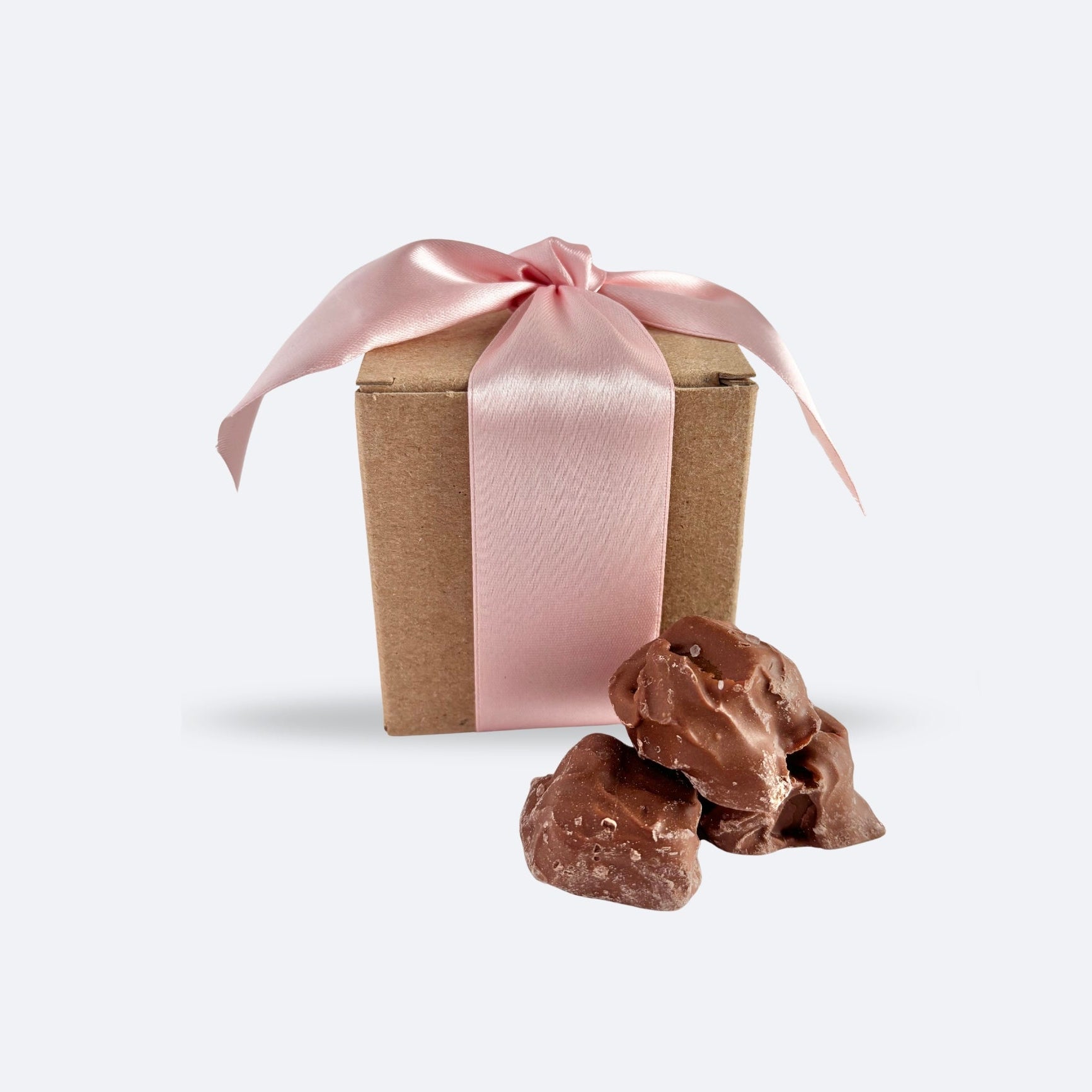 Chocolate sea salt caramels are featured in image inside of kraft box with pink ribbon with 3 caramels outside on a white background. 