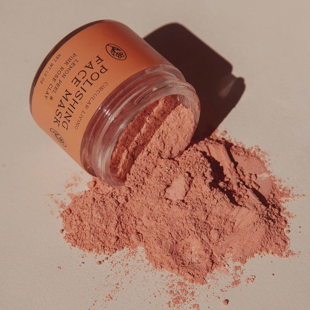 A clear jar of Polishing Face Mask powder lies on its side with the pinkish-terracotta powder spilled out onto a neutral beige surface. The brand logo 'The Body Shop' is visible on the label.