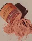 A clear jar of Polishing Face Mask powder lies on its side with the pinkish-terracotta powder spilled out onto a neutral beige surface. The brand logo 'The Body Shop' is visible on the label.
