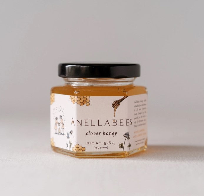 clover Honey form Anellebees, picture of it's container transparent hexagon shaped jar with white label that has images of honey combs and a honey drizzle pouring a bit of honey. The jar shows the honey inside in its yellowish caramel color.  