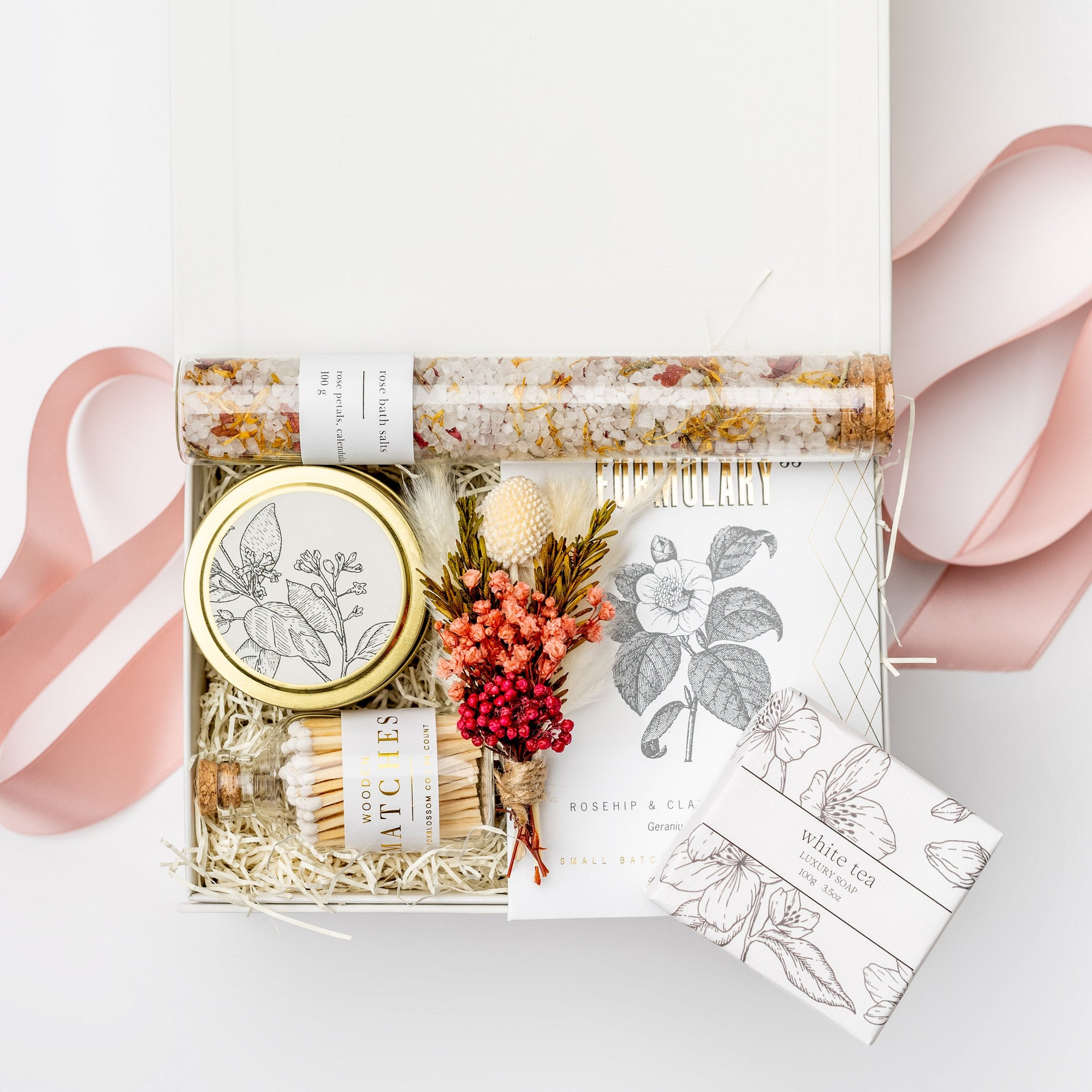 A top view of the Coming Up Roses Spa Gift Box featuring,  a 4 oz. hand-poured Santal & Vanilla candle, a white matchsticks jar, a Formulary 55 Rosehip & Clay facial mask, a white tea shea butter guest soap, rose bath soaking salts, and a handmade dried floral mini bouquet. Arranged in a white gift box filled with ivory shred and wrapped with a soft pink ribbon around the box on a white background.