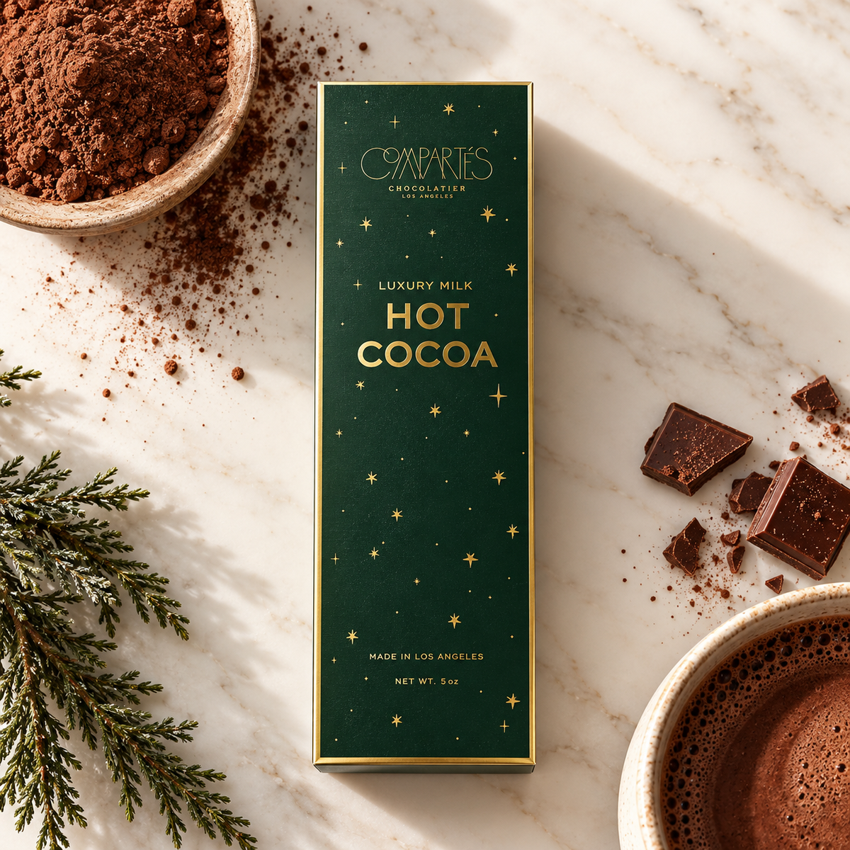 A tall rectangular box of Compacts Chocolate Company Luxury Milk Hot Cocoa sits on a white marble surface with cocoa powder, chocolate chunks, evergreen branches, and a cup of hot cocoa arranged around it.