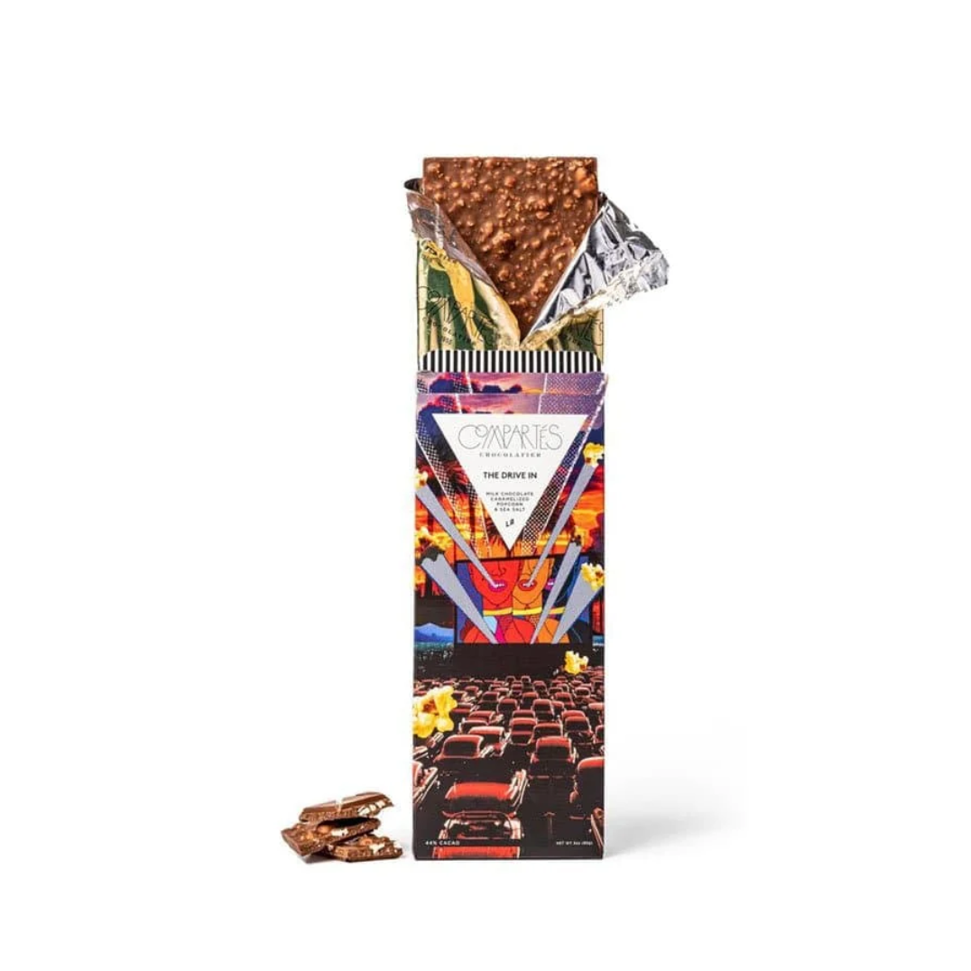 The drive in Compartes' chocolate bar is feautured,  a chocolate bar, partially unwrapped to display its contents. The chocolate has a textured surface, possibly due to inclusions like nuts or crispy rice. The packaging is a key element, featuring a design with the "Compartes" logo prominently displayed, along with graphics related to "The Drive In." These graphics likely depict a movie scene, using vibrant colors, with a mix of text and imagery to catch the eye on a white background. 