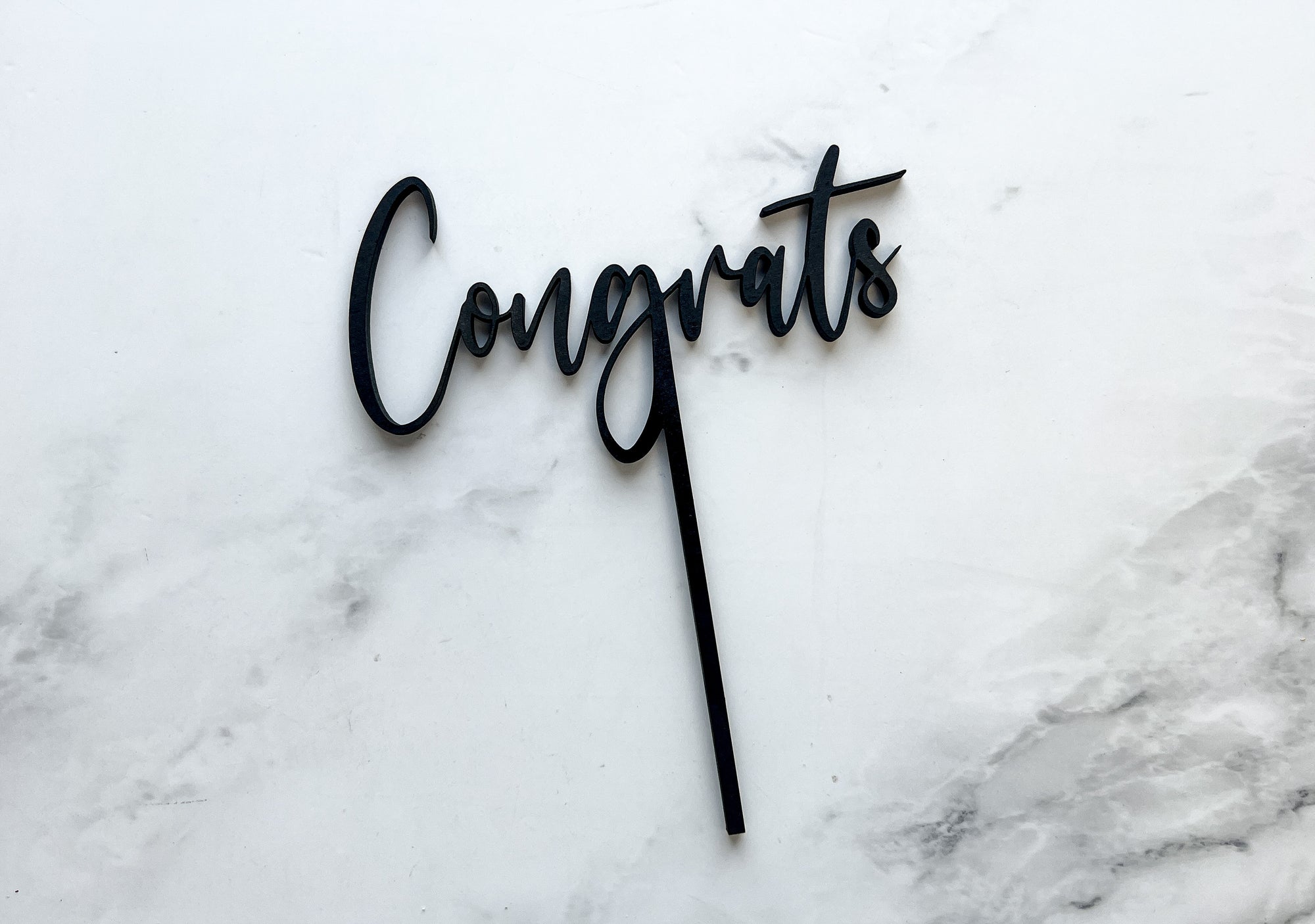 Congrats wooden cake topper is featured in matte black on a marble like background. 