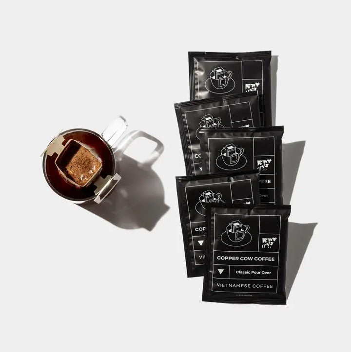  Copper Cow Vietnamese Coffee, mug with a metallic pour-over filter on the left and four packets of the Vietnamese coffee in its black sachets with white lettering, all arranged on a clean white surface.