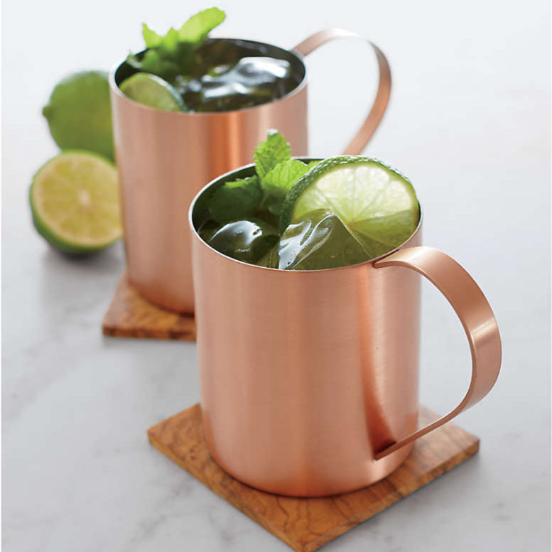 Two copper mugs filled with the Moscow Mule drink, garnished with lime wedges and mint leaves, sit on wooden coasters. With twolimes in the background which is white. 