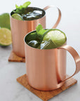 Two copper mugs filled with the Moscow Mule drink, garnished with lime wedges and mint leaves, sit on wooden coasters. With twolimes in the background which is white. 
