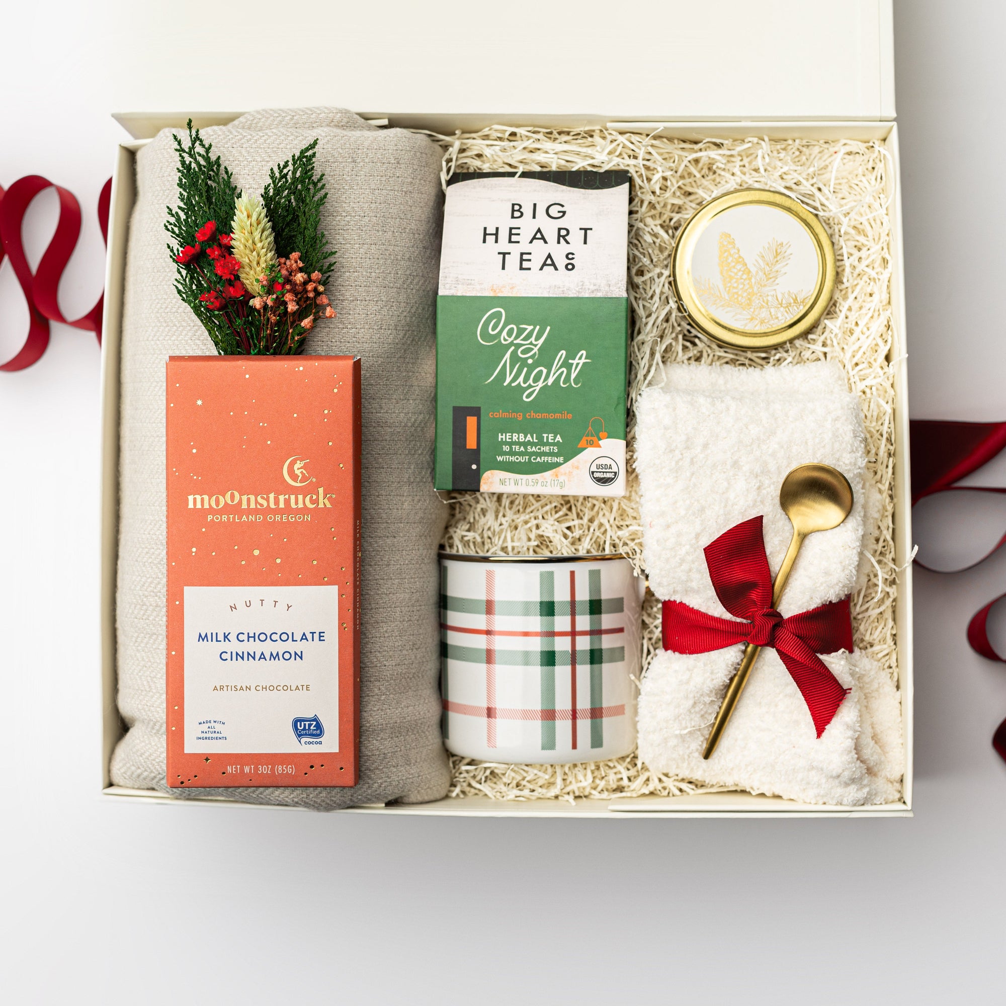Cozy at home holiday gift box is featured  in a white box with ivory shred on the back with red ribbon underneath it.  On the left gray throw blanket with a moonstruck nutty milk chocolate cinnamon chocolate bar, and mini dried floral bundle. In the middle cozy night herbal tea, plaid mug green and red stripes. On the right spruce gold candle, white cloud socks tied in red ribbon and gold platted stirring spoon. On a white background. 