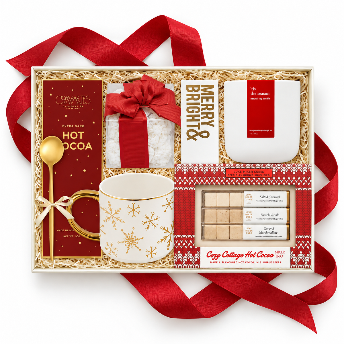 A holiday hot cocoa gift set in a white box filled with shredded paper, featuring a white ceramic mug with gold snowflakes, a red and white hot cocoa box, a white candle, a gold spoon, and chocolate bars, all arranged with a red ribbon on a white background.