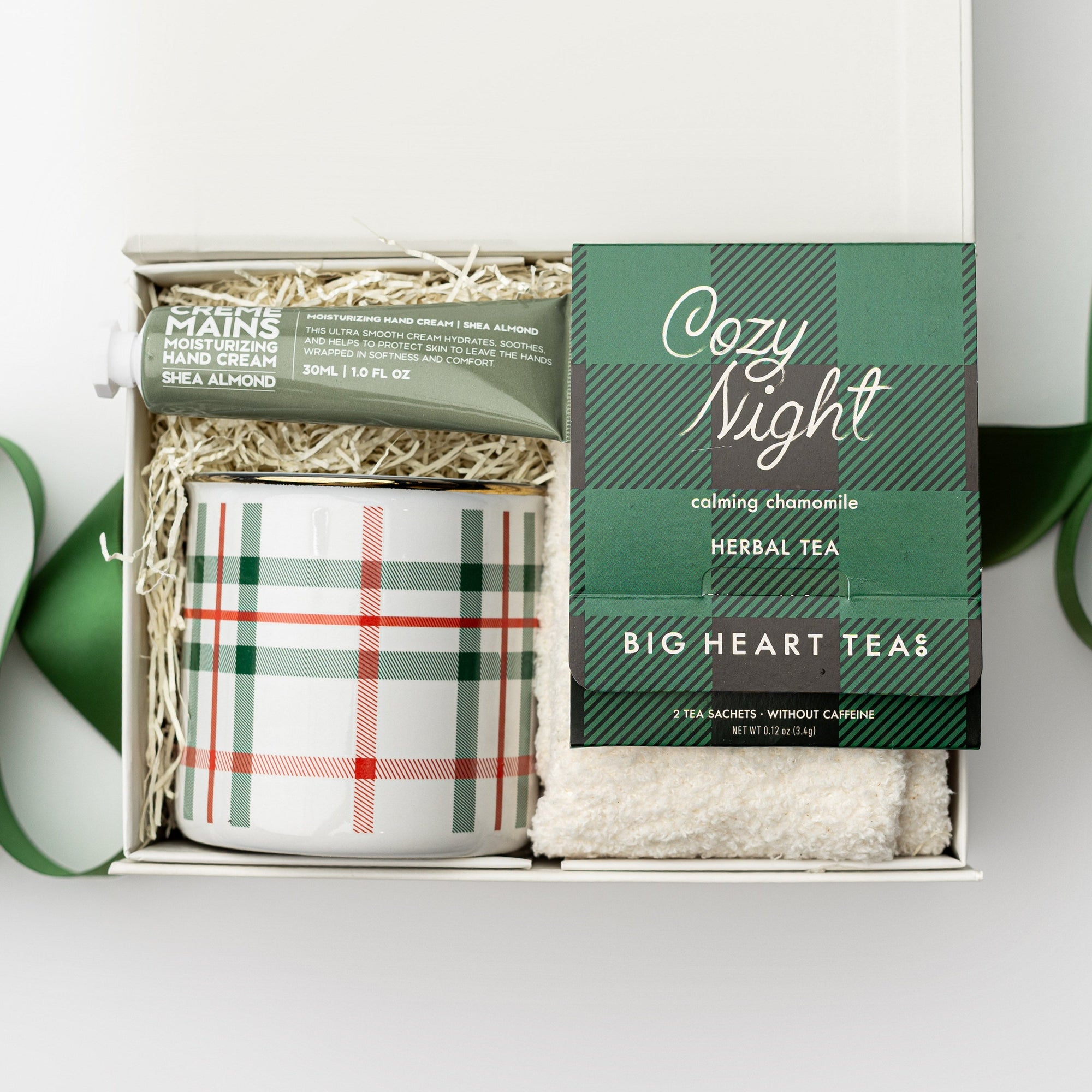 Petite cozy at home gift box featured in a small white box with ivory shred in the back with green ribbon crossing under it. Inside of the box on the left side almond and shea butter hand cream in its green tube container and plaid ceramic mug with white and green lines. On the right cozy night herbal tea in its green and black  plaid pattern package and underneath white cozy socks. All on a white background. 