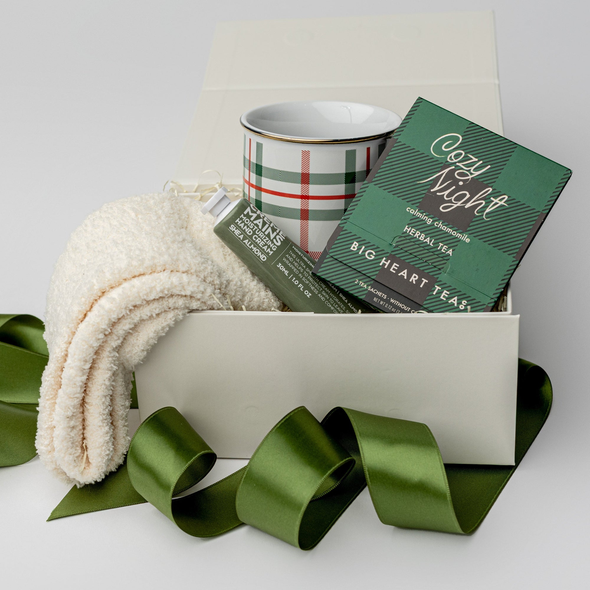 Cozy Night Holiday Gift is featured in a white box wrapped with a green ribbon. Inside the box, on the right, are white cozy socks; in the middle, a plaid mug with green and red lines; and on the left, a Cozy Night herbal tea in black and green plaid packaging. Everything is displayed on a white background.