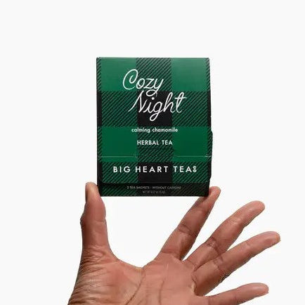 Cozy Night Tea presented in its green and black plaid package and, white lettering, Being held by an african-american hand on a white background. 