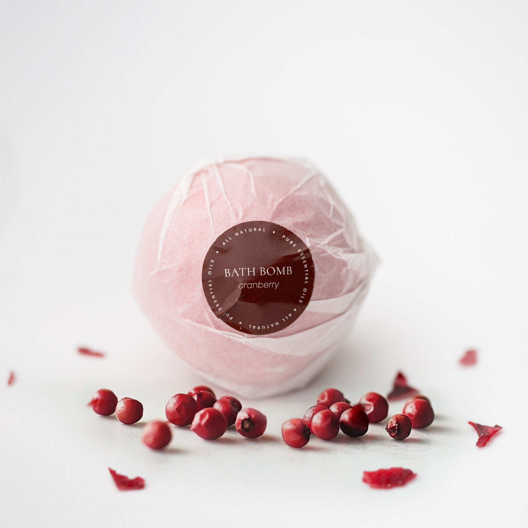 Cranberry bath bomb is featured in its white transparent covering with red label that has white lettering., Surrounded by cranberries on a white background. 