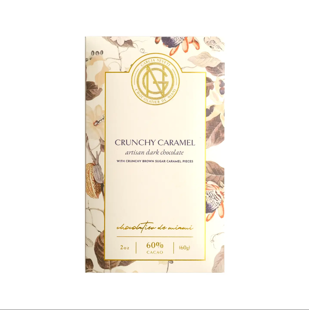 Crunchy caramel dark chocolate bar is featured in its rectangular-shaped package with cacao tree illustration in brown, grey, and beige colors with a white label with gold rim foil printed on it with gray and gold lettering on a white background.
