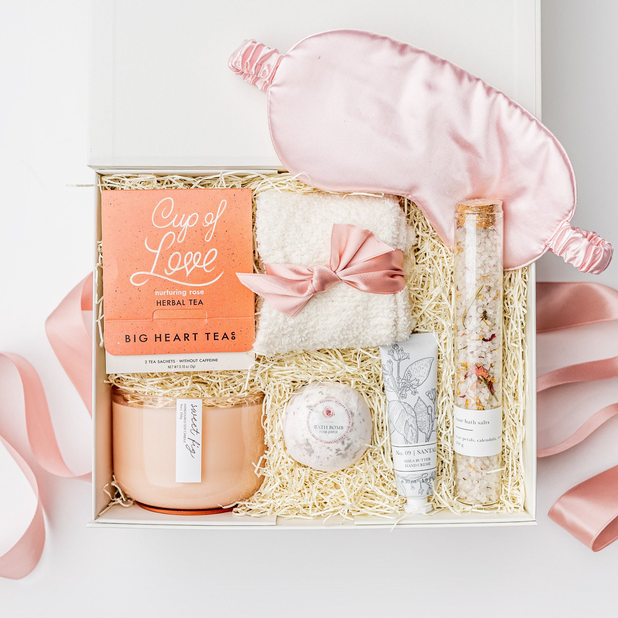 A white gift box filled with pink ribbon and ivory paper shred on a white background. Inside the box are a pink satin sleep mask, Cup of Love rose tea for two, an 11oz hand-poured soy double-wick candle in a keepsake glass jar labeled “Sweet Fig,” plush ivory cloud socks, a tube of shea butter hand crème in the scent Santal Vanille, a rose petal bath bomb, and a jar of rose bath salts — a cozy, self-care “Cup of Love” gift box.