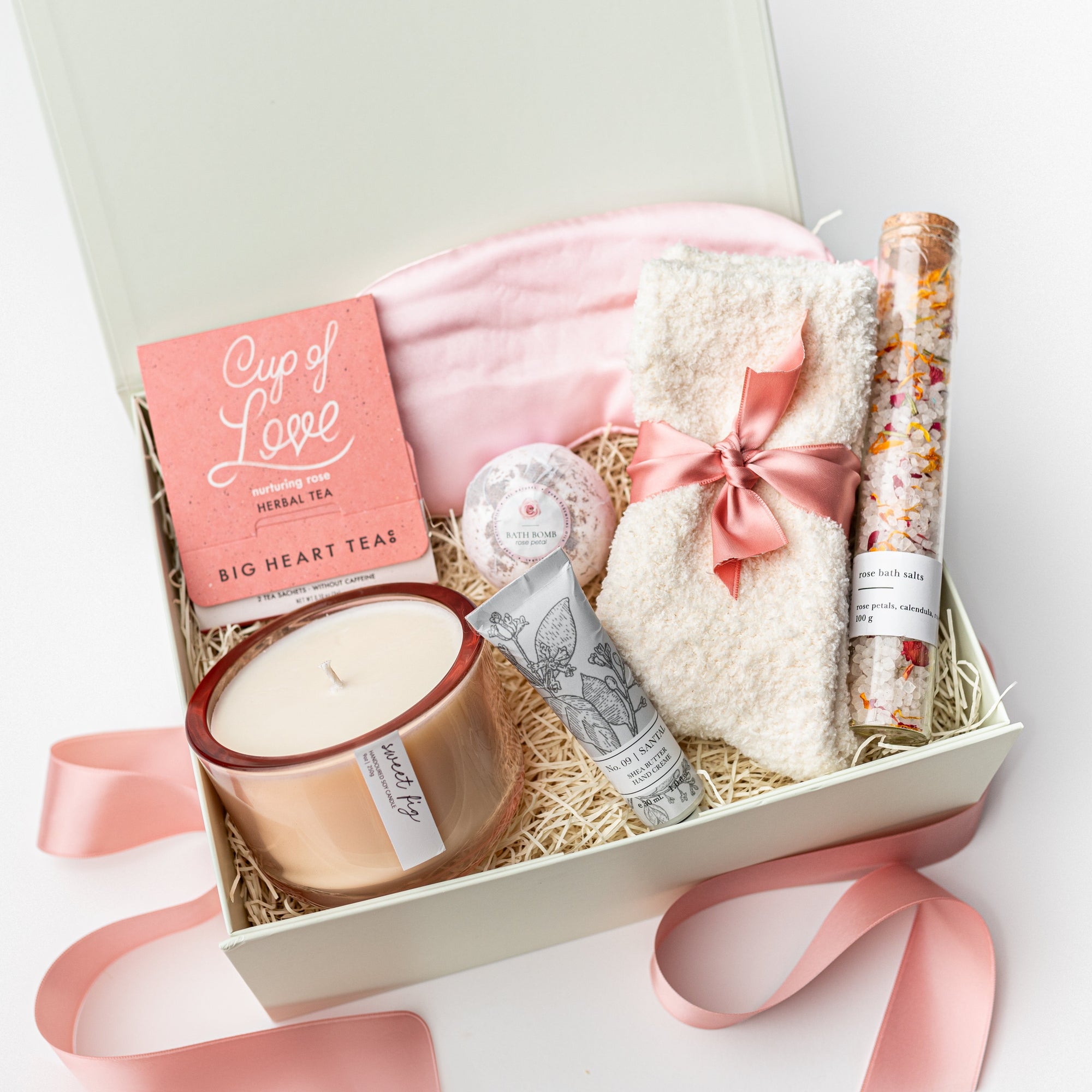 A top view of a luxurious self-care gift set featuring a pink satin sleep mask, a Cup of Love rose tea for two, a 9 oz. Sweet Fig luxury keepsake candle, cloud ivory socks, No. 09 Santal shea butter hand crème, a rose petal bath bomb, and rose bath salts, all arranged in a white gift box with ivory shred and tied with a soft pink ribbon on a white background.